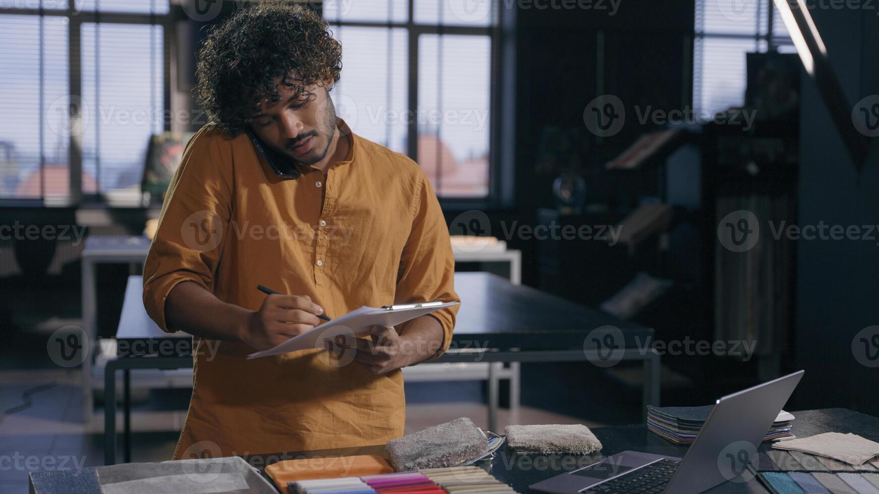 Arabian Indian male business man working busy interior designer talking with client online discuss on mobile phone fabric tissue samples write in notepad textile order in workshop modern design studio photo