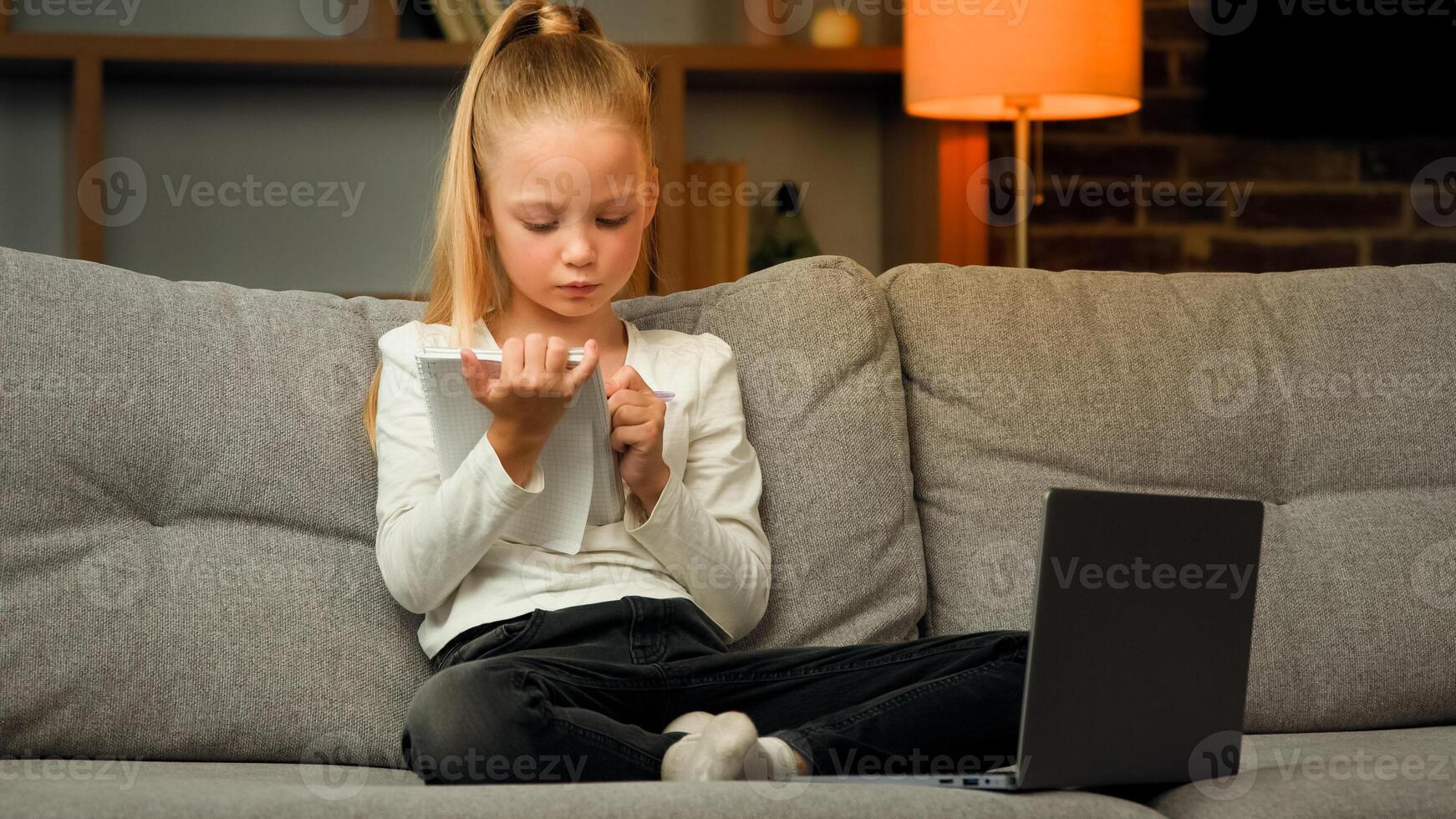 Concentrated pupil girl making homework at copybook on cosy sofa writing math tasks of elementary school subjects practicing calligraphic handwriting learn language by online educational lesson photo