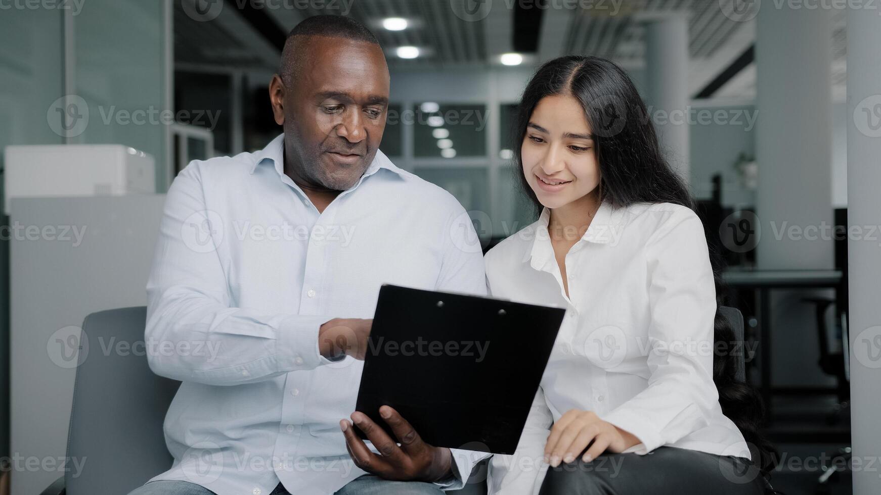 Diverse business partners sit in office discuss paper instruction african businessman show documentation in folder tablet to arabian female colleague explain insurance contract benefits negotiating photo