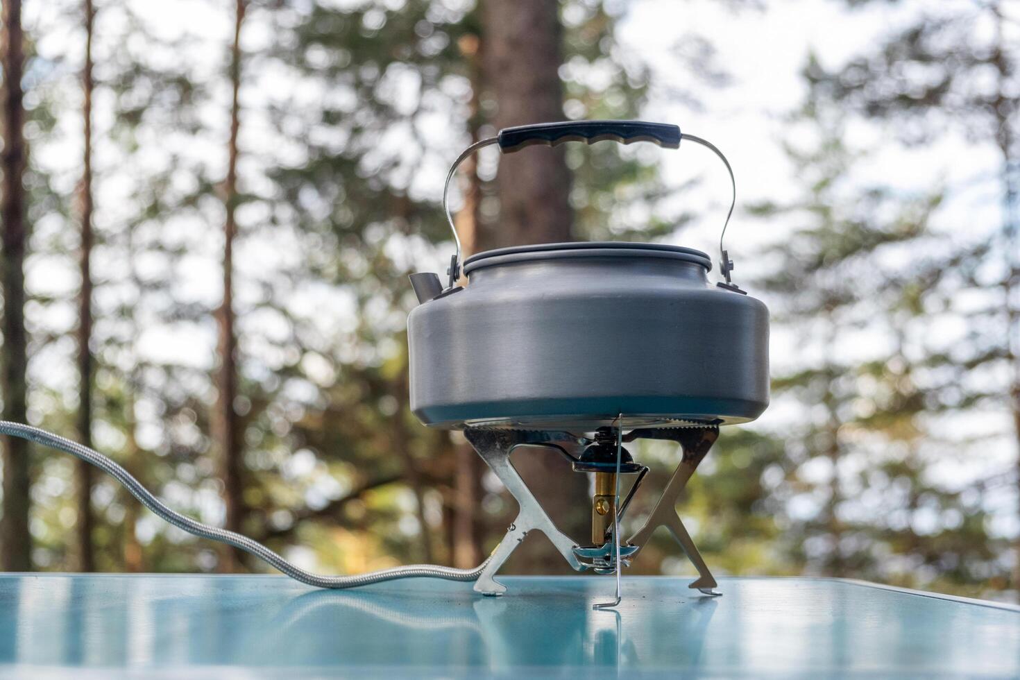 A kettle heats water on a small camping stove in a natural forest environment under clear skies in the afternoon photo