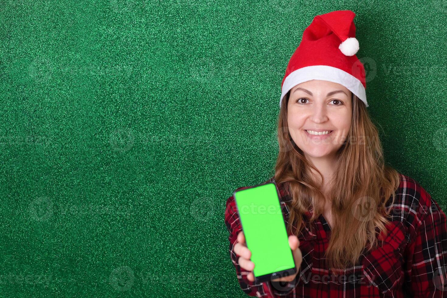 beautiful girl in a New Year's cap on a green background shows a phone screen with a chromakey background. Christmas. New Year photo