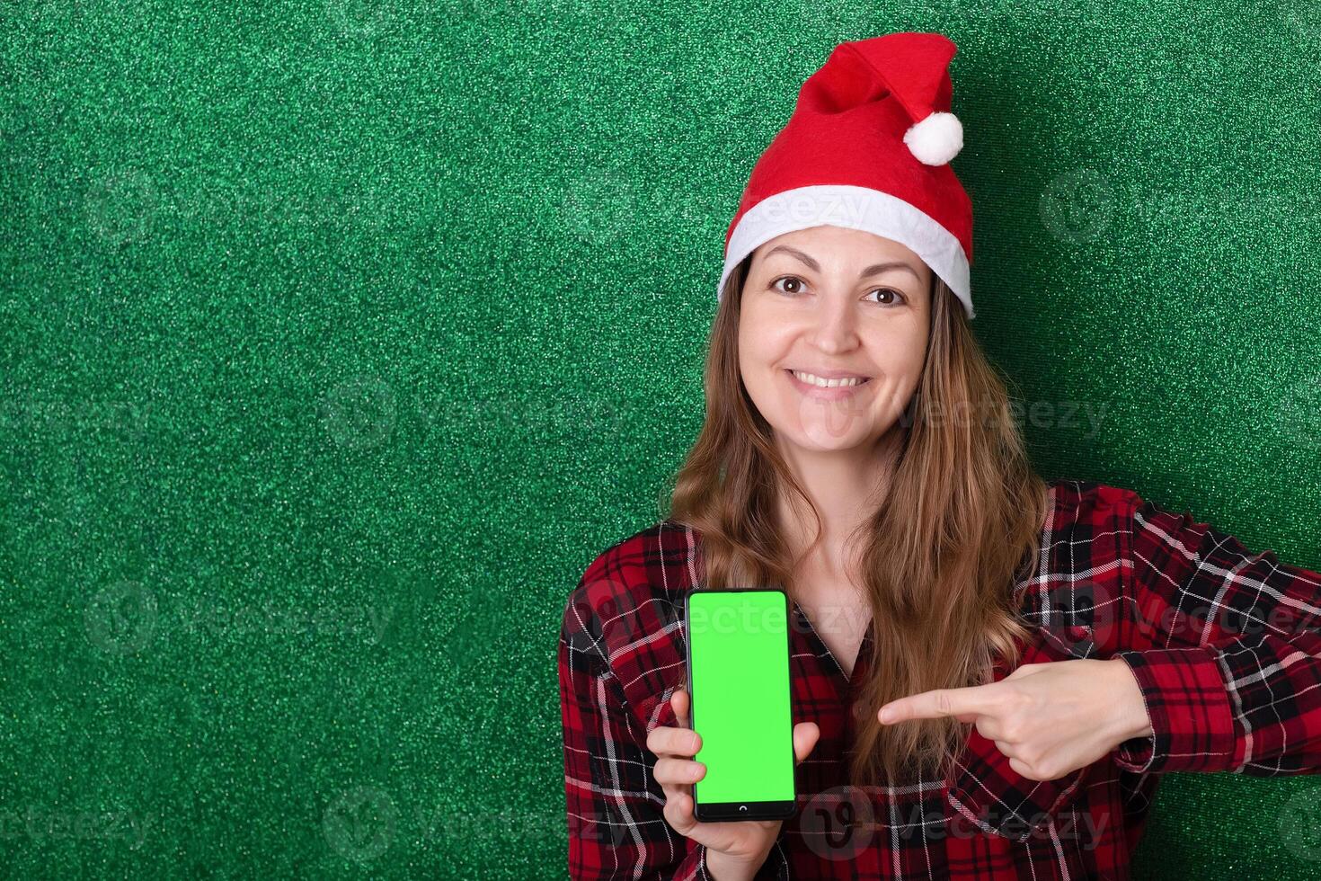 beautiful girl in a New Year's cap on a green background shows a phone screen with a chromakey background. Christmas. New Year. photo