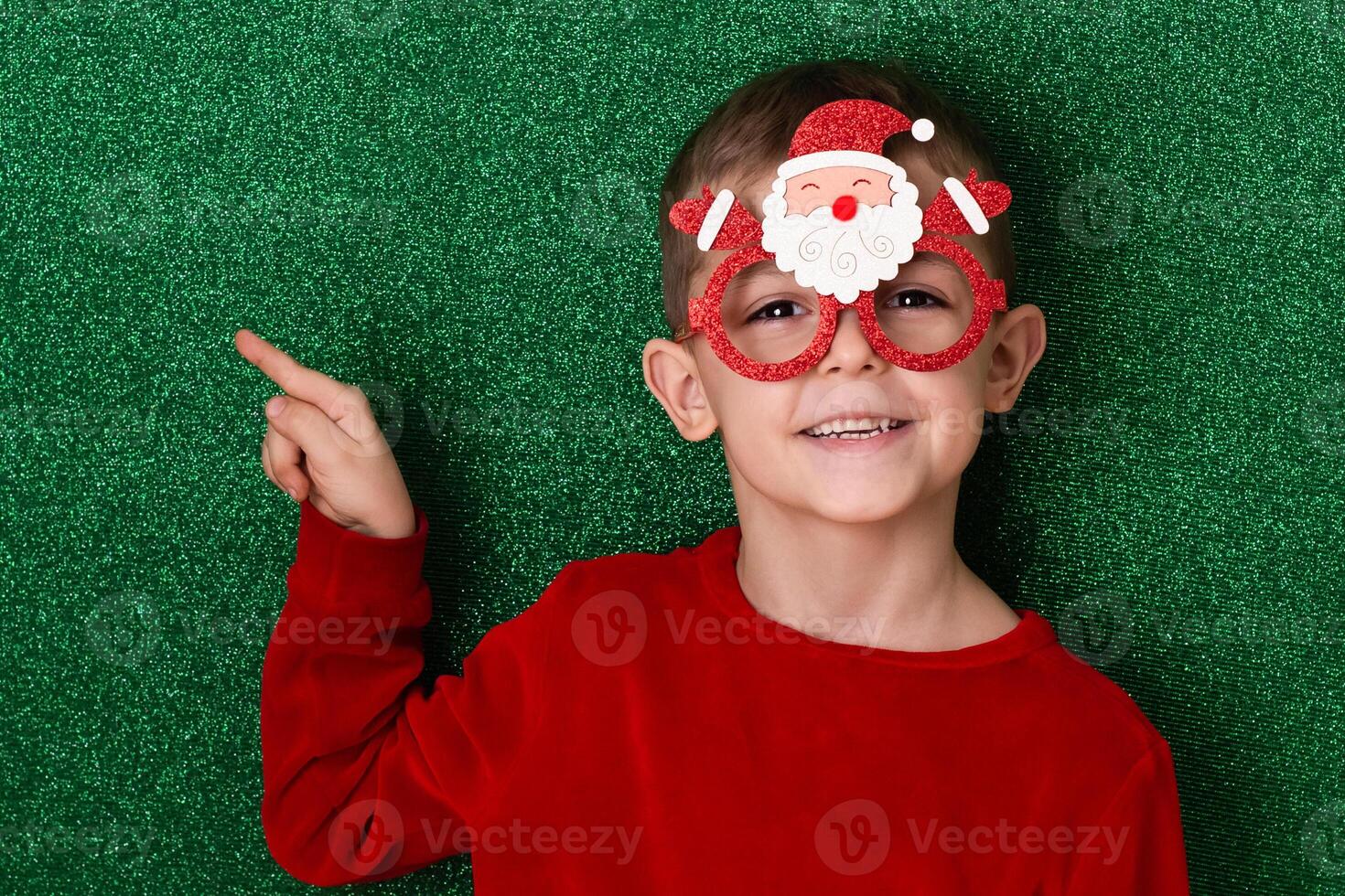 a cute boy in New Year's souvenir glasses with Santa Claus on a green background points to the background. New Year photo