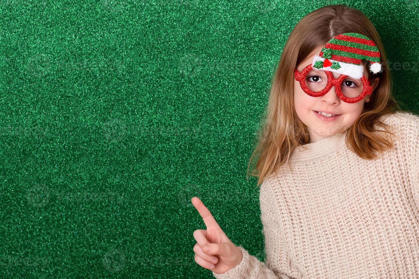 a cute girl in New Year's souvenir glasses with a New Year's cap on a green background points to the background. New Year photo
