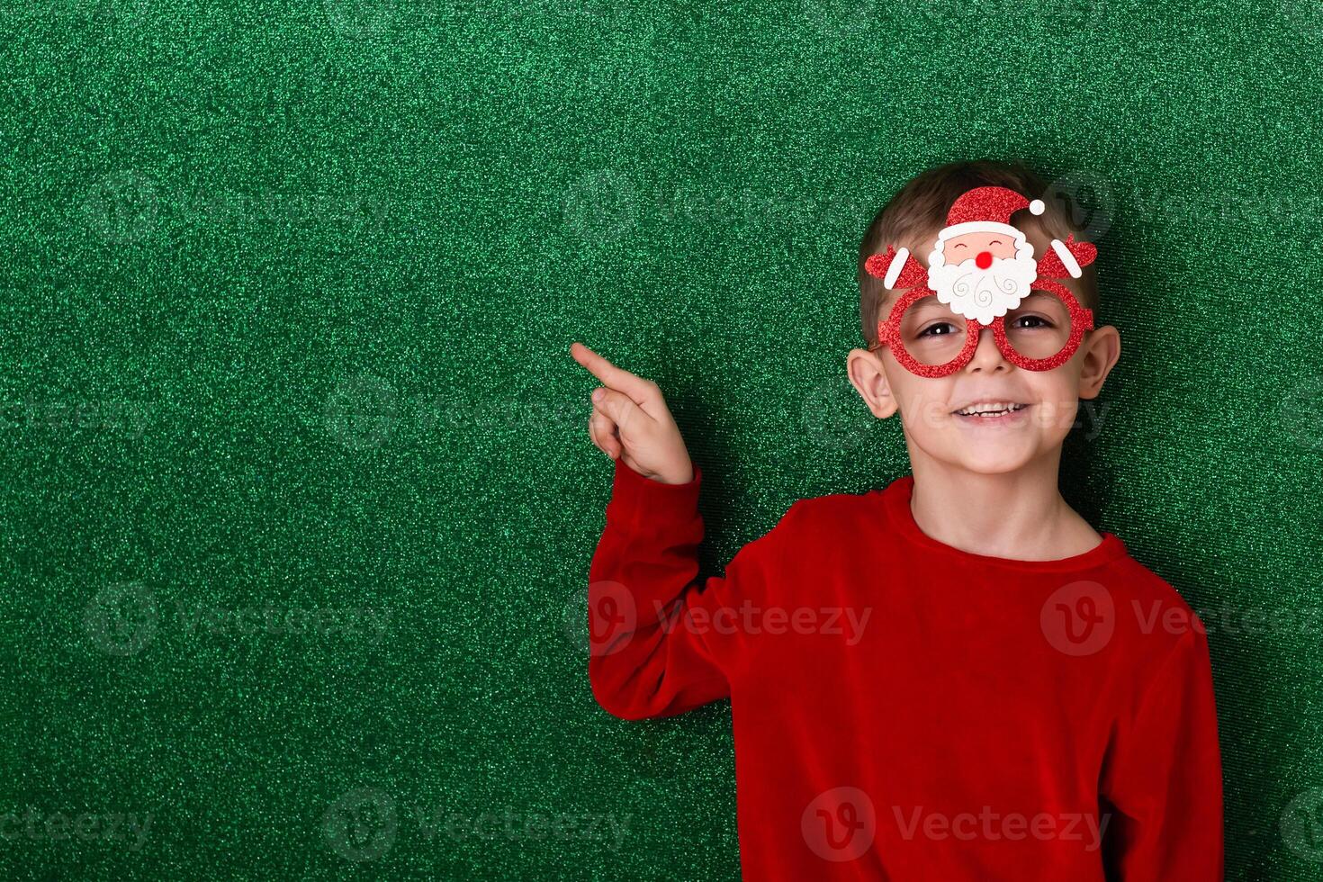 a cute boy in New Year's souvenir glasses with Santa Claus on a green background points to the background. New Year photo