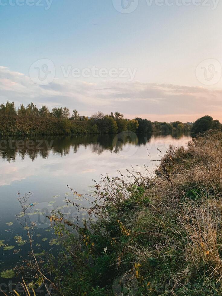 Serene river flows peacefully under soft sunset sky. Lush greenery with wild grasses and plants. Decorative landscape, design element for background about nature, fresh and beauty of environment photo