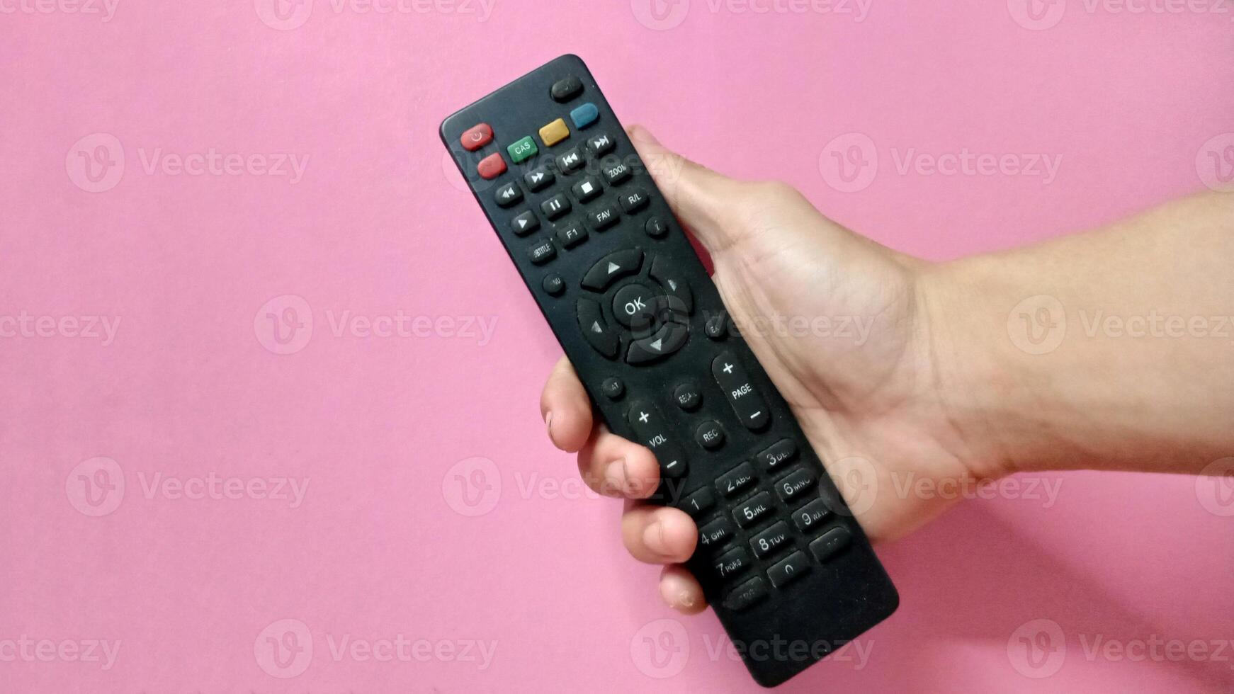 Close up of a hand holding a black TV remote control against a pink background. Concept of technology, entertainment, and media. photo