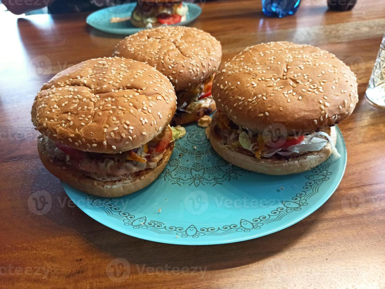 Three homemade hamburgers sit on a decorative blue plate. Each burger features a sesame seed bun, grilled meat, and an assortment of fresh vegetables, embodying a delightful meal experience photo