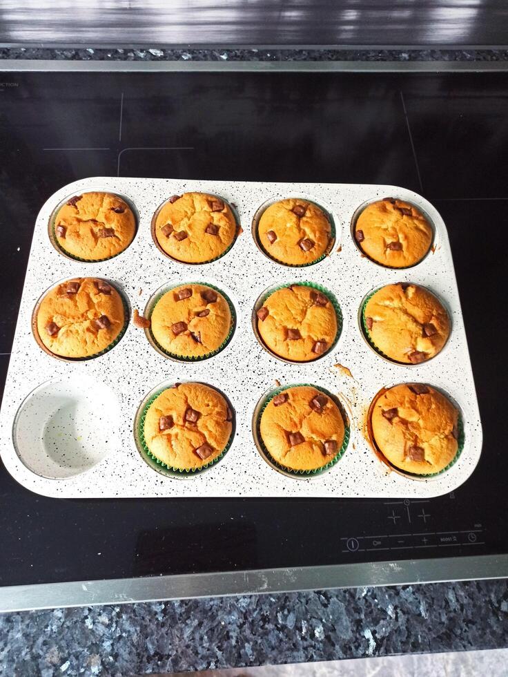 A muffin tray filled with batter and chocolate chunks sits on a kitchen counter, ready to be baked. The baking cups are lined and each has a generous amount of chocolate photo