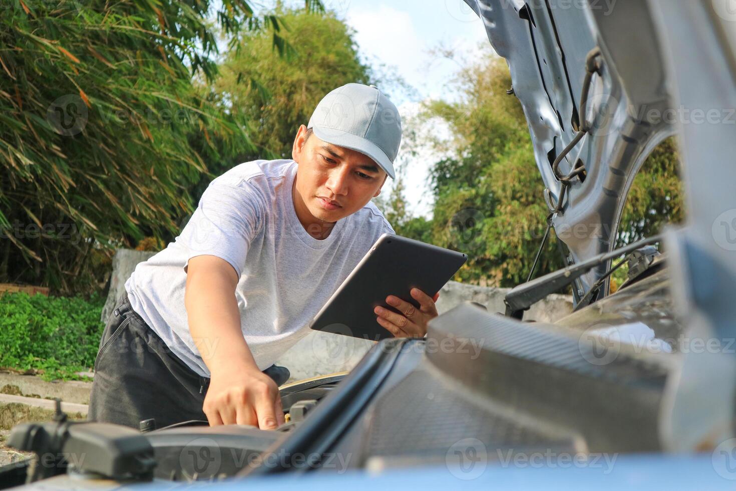 A young mechanic opens the hood of a car to check for engine damage and perform professional maintenance. He wears a grey uniform and inspects and repairs his car in the maintenance center. photo