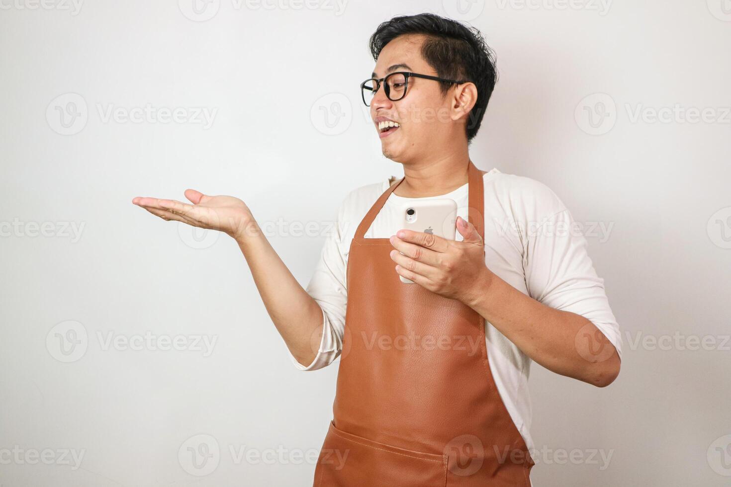 Excited young asian man wearing rolled-up white sleeve shirt and brown apron is opening hands receiving something with smiling face expression. Isolated over white background photo