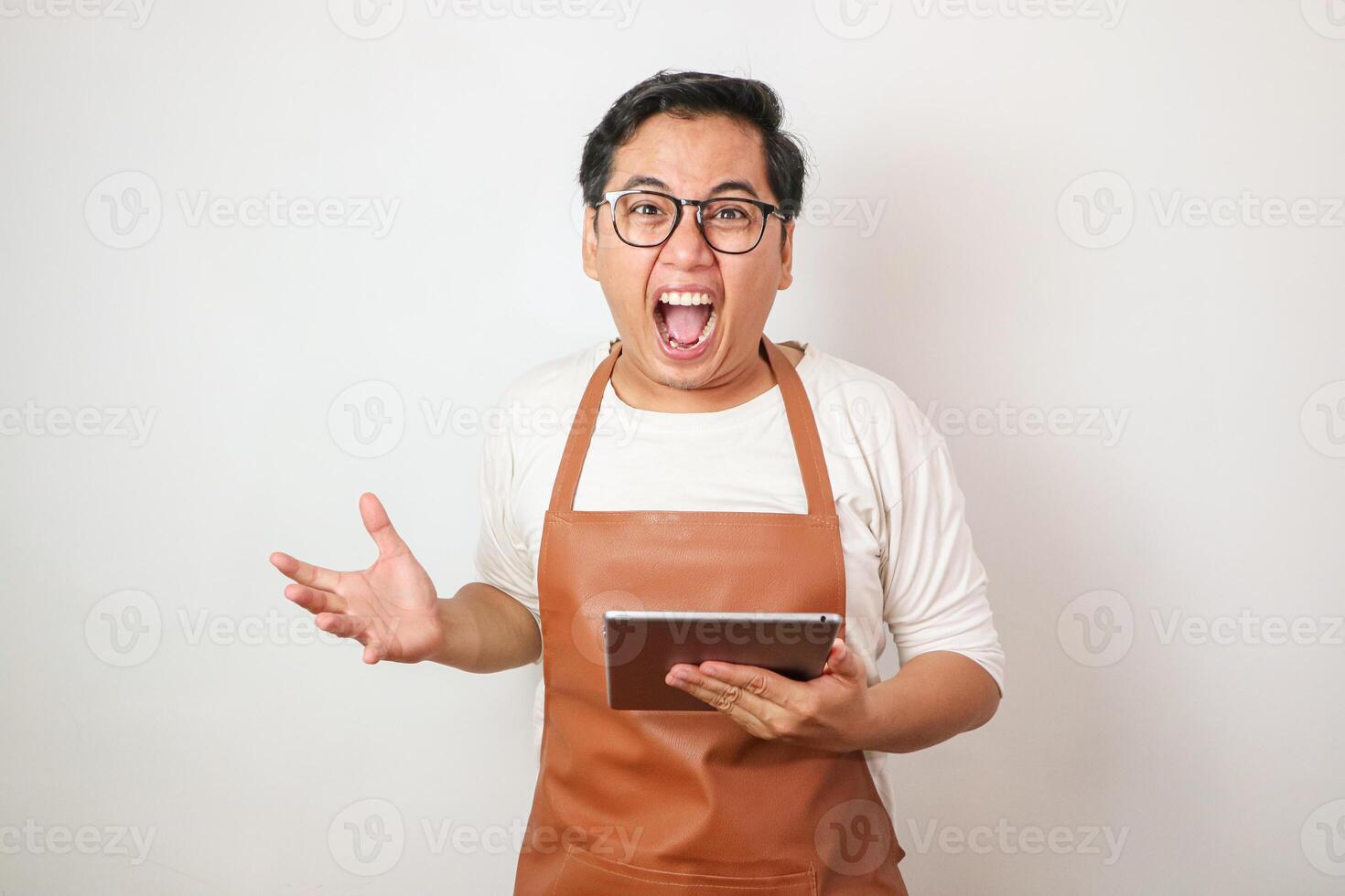Asian waiter man in apron holding tablet mad while yelling with aggressive expression and arms raised. photo
