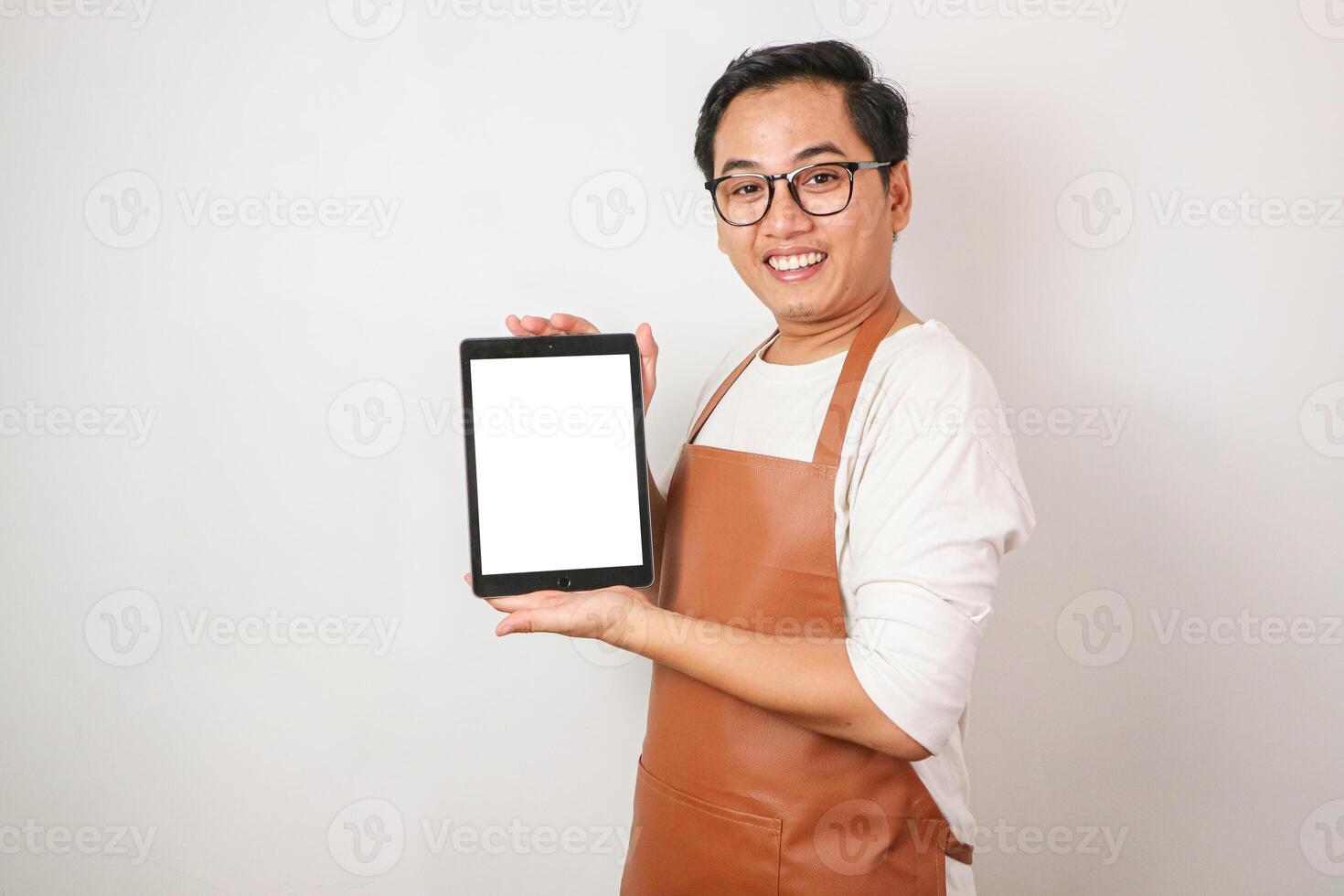 smiling young Asian man barista barman employee wearing brown apron work in coffee shop holding white blank tablet and looking at copy space isolated on white background. photo