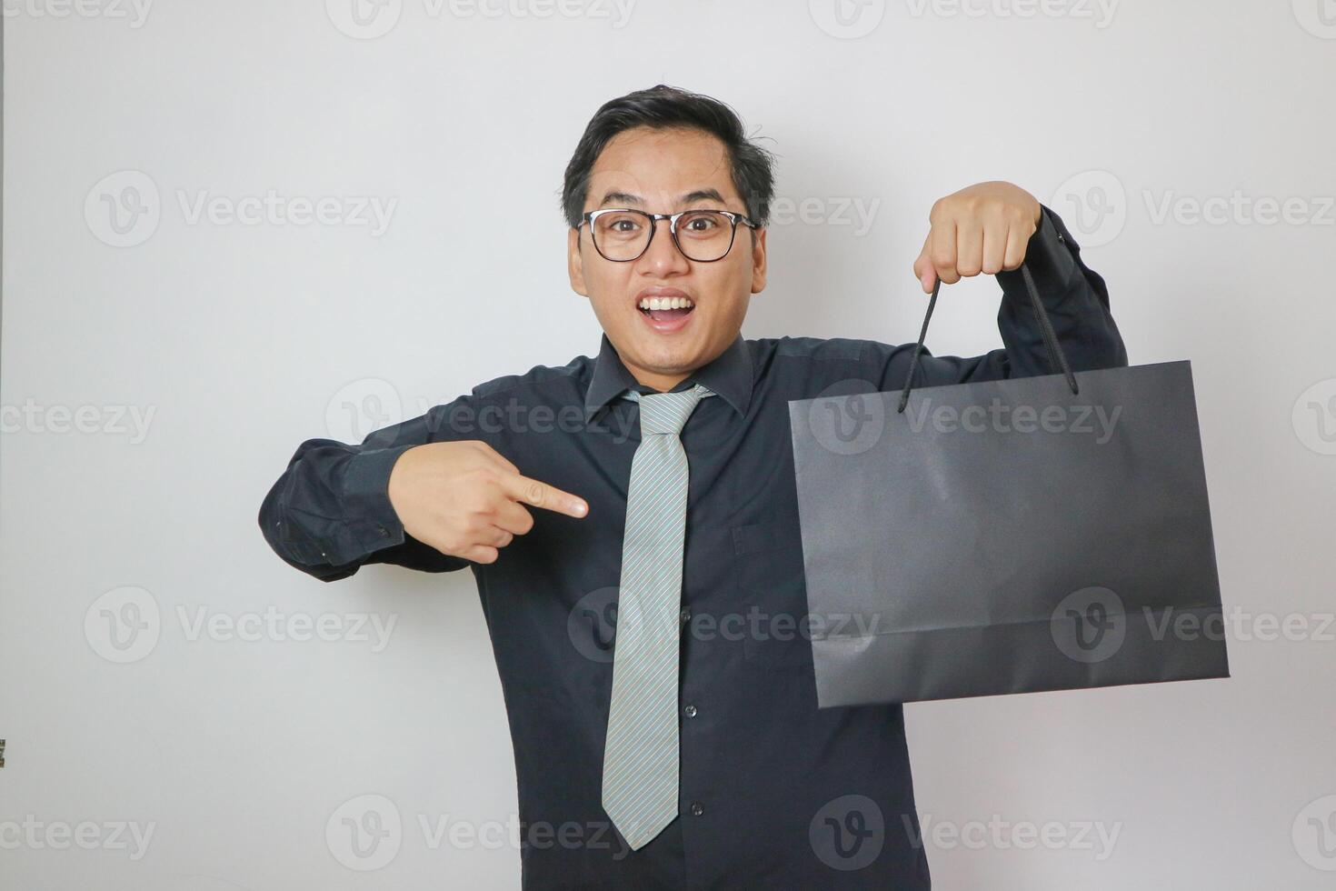 Asian office worker wearing black shirt pointing at black shopping bag, isolated white office worker concept. photo
