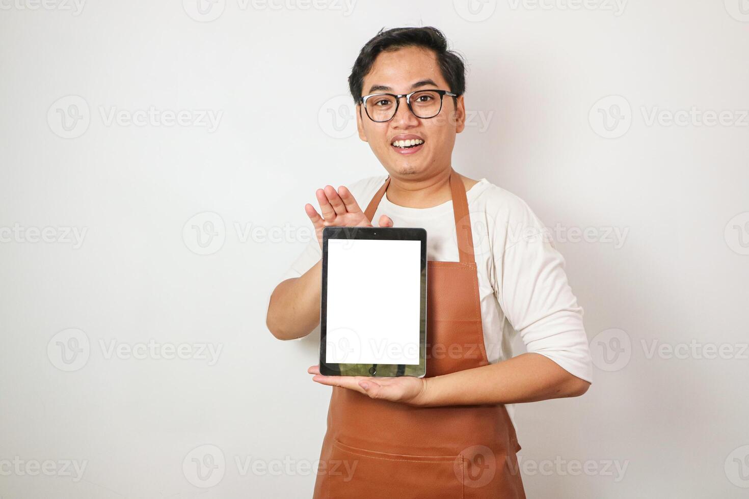 Excited young asian man wearing rolled-up white sleeve shirt and brown apron is showing vertical blank white screen tablet with smiling face expression. Isolated over white background. photo