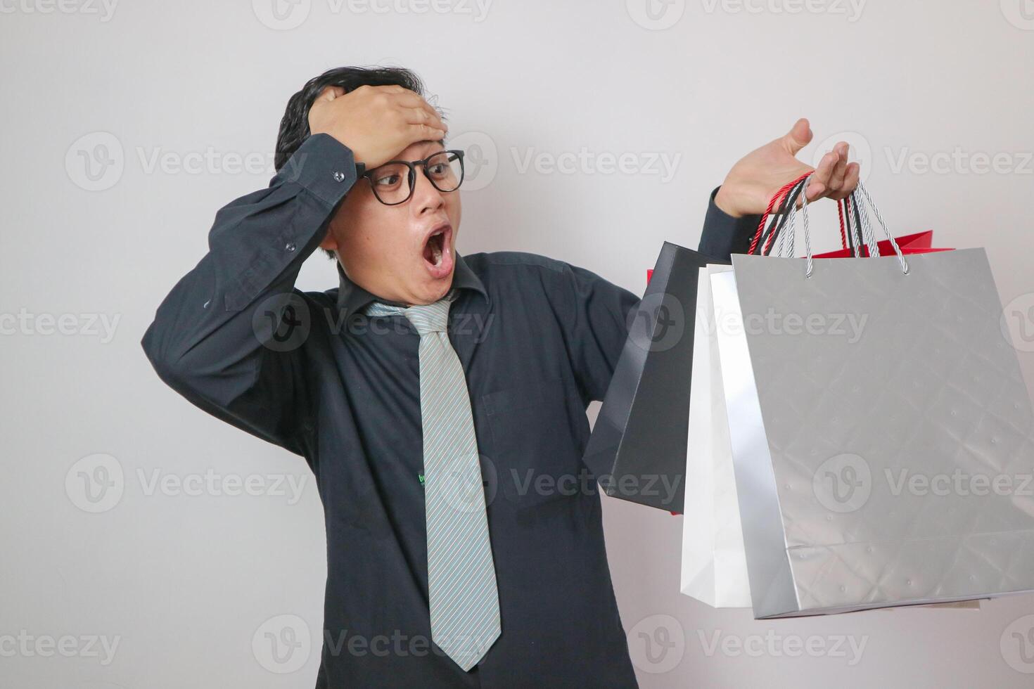 Portrait of a surprised Asian man wearing a black shirt holding shopping bags, looking at the camera with a surprised and astonished facial expression. Indoor studio shot isolated on blue background. photo