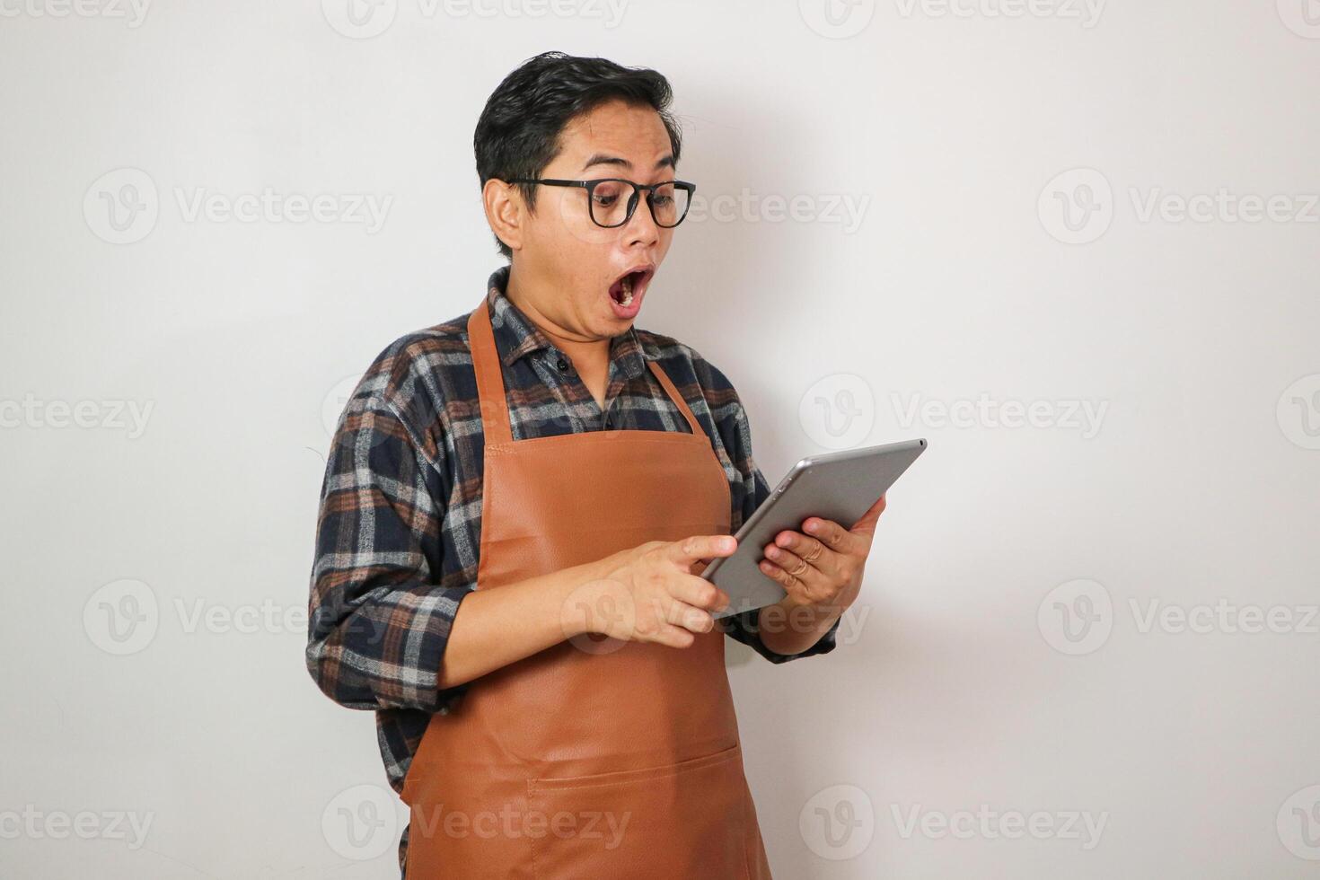 asian man wearing apron holding tablet with surprised expression looking at tablet, coffee and restaurant business idea concept photo