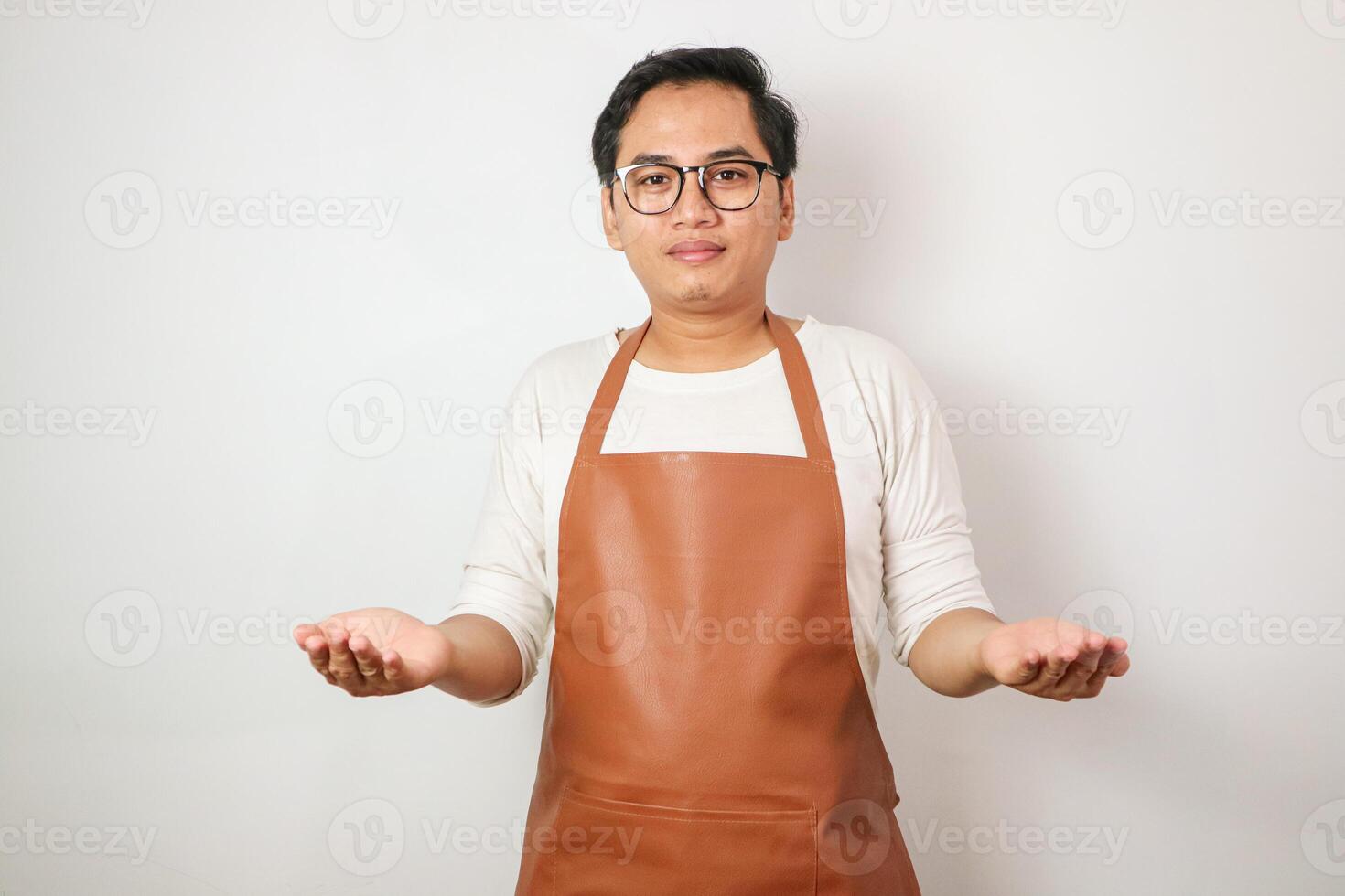 Excited young asian man wearing rolled-up white sleeve shirt and brown apron is opening hands receiving something with smiling face expression. Isolated over white background photo