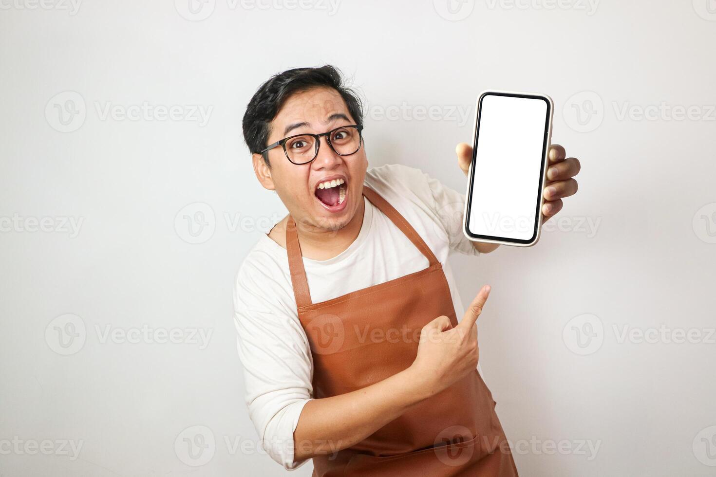 Shocked young asian man wearing rolled-up white sleeve shirt and brown apron is holding a smartphone with surprised face expression. Isolated over white background. photo