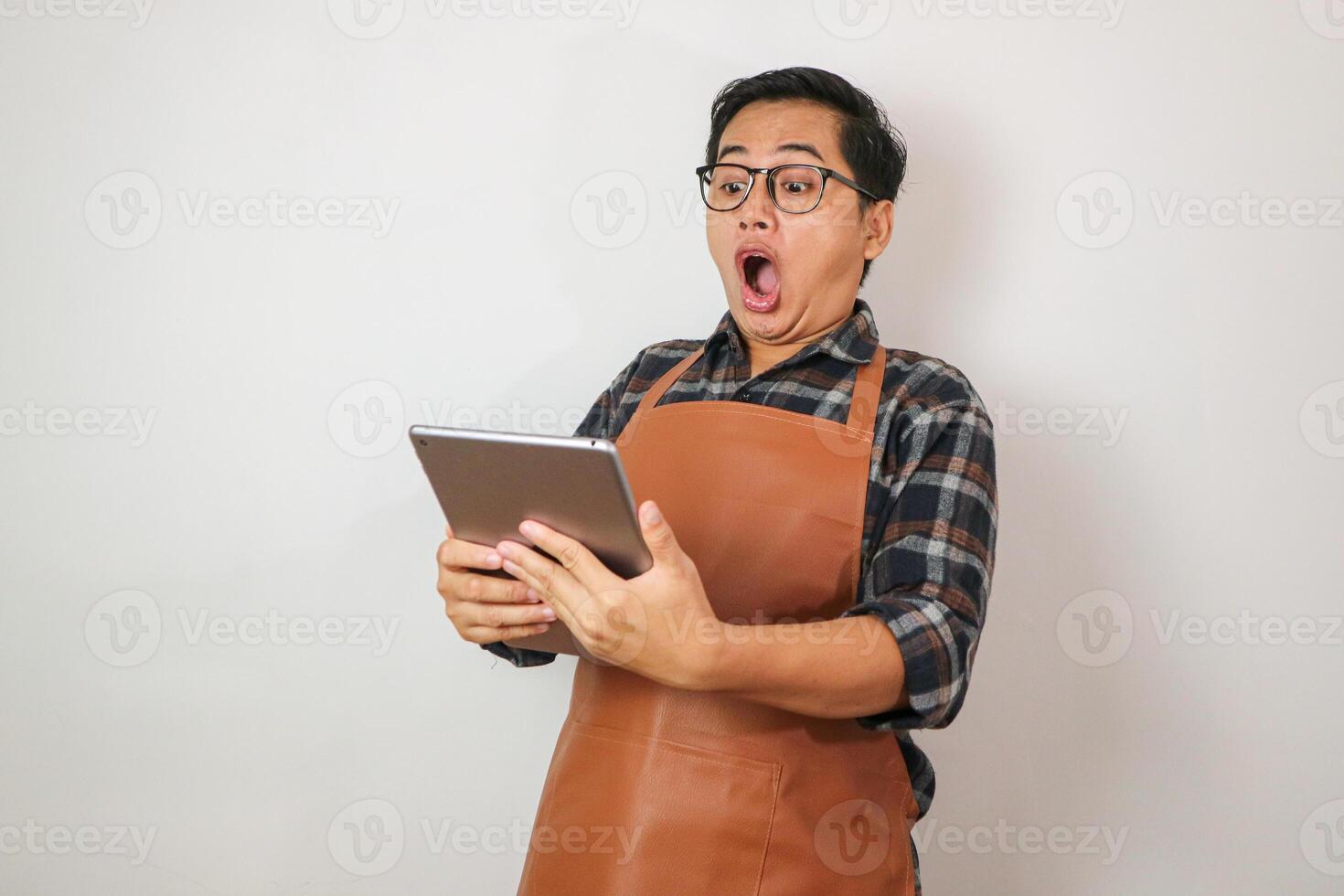 Asian man wearing apron holding tablet with surprised expression looking at tablet, coffee and restaurant business idea concept photo