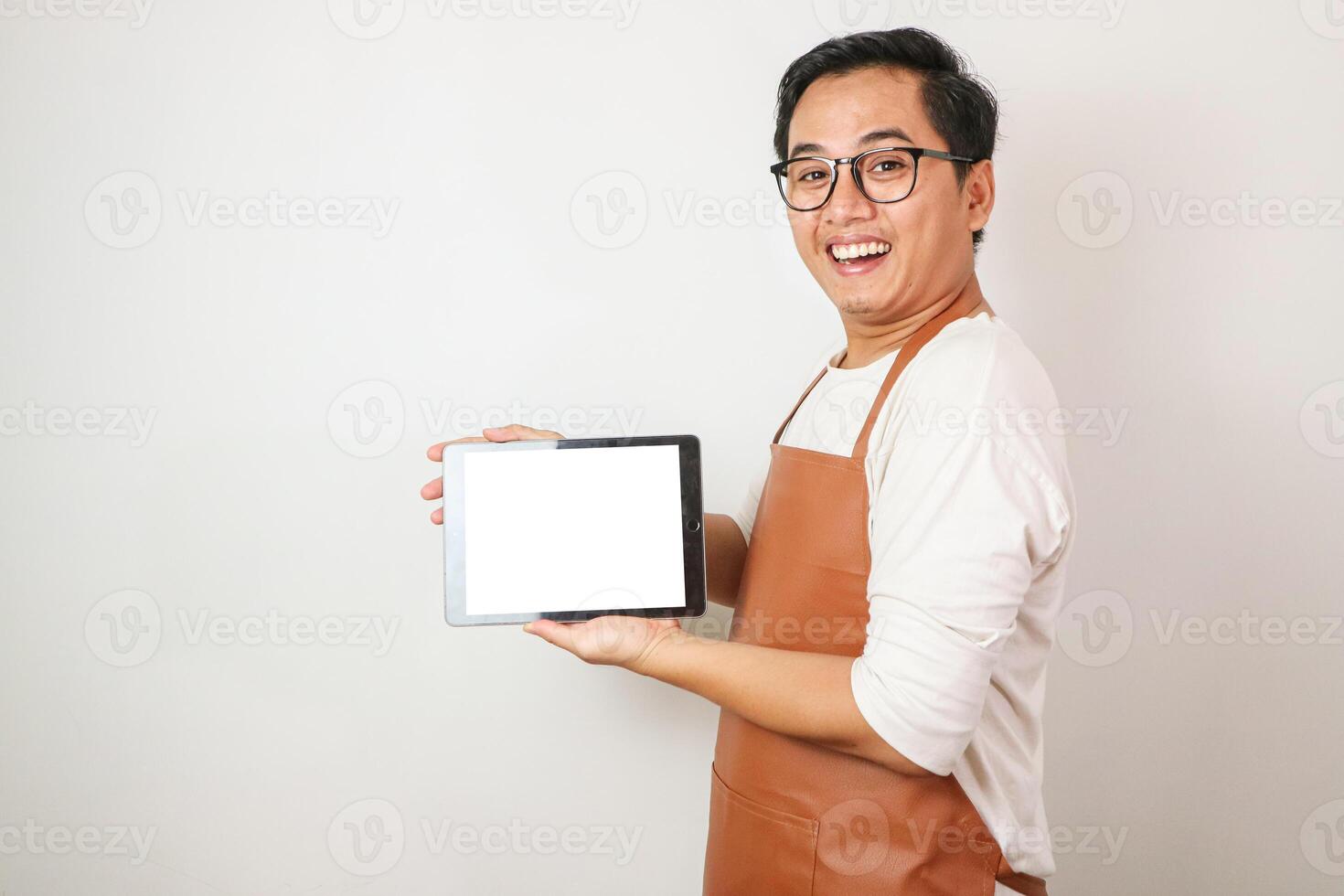Excited young asian man wearing rolled-up white sleeve shirt and brown apron is showing vertical blank white screen tablet with smiling face expression. Isolated over white background. photo