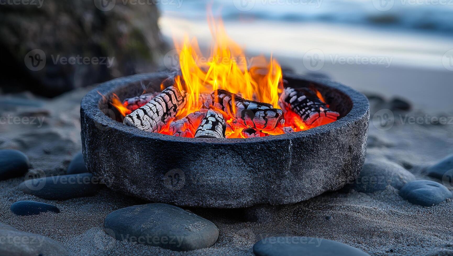A fire is burning in a large bowl on a beach. The fire is orange and yellow, and the bowl is made of stone. The scene is peaceful and relaxing, with the sound of waves in the background photo