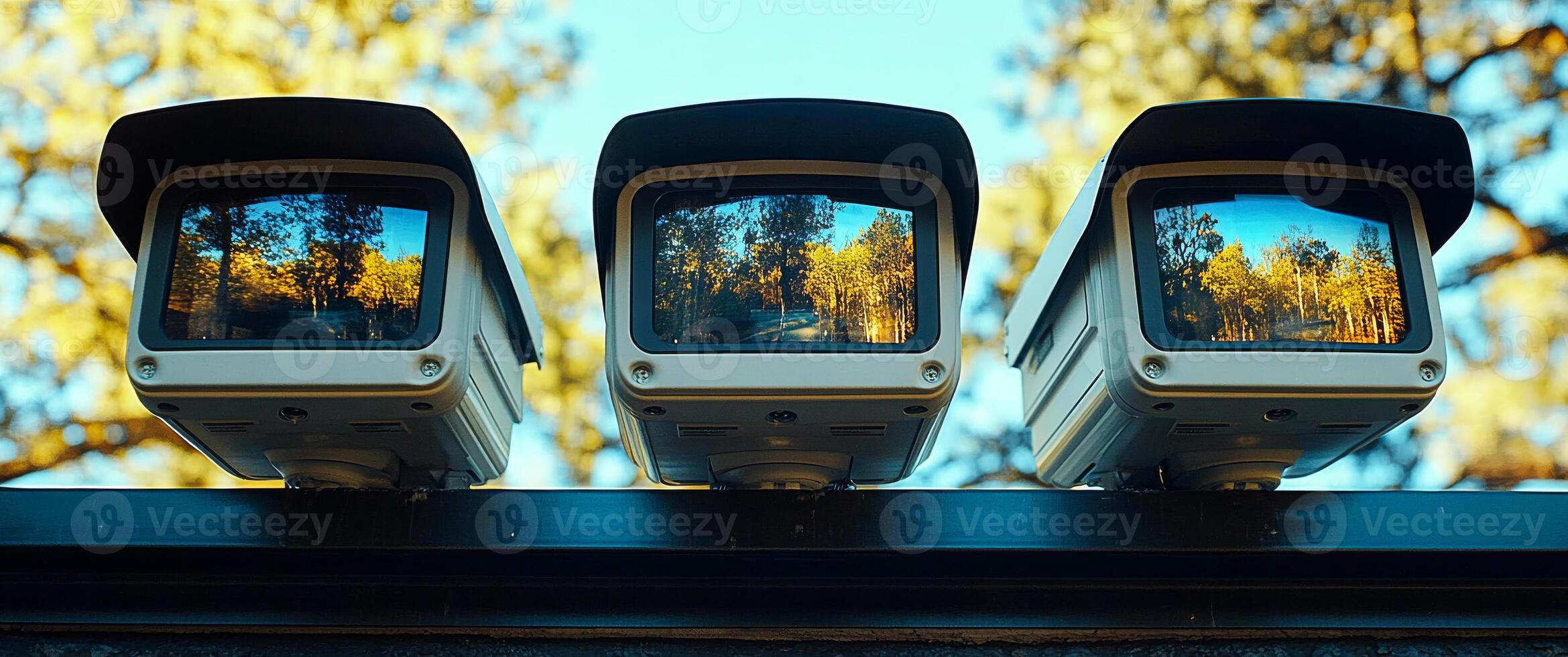 Security cameras capturing serene outdoor views. Three security cameras lined up, displaying vibrant autumn trees and a clear sky in the background. photo