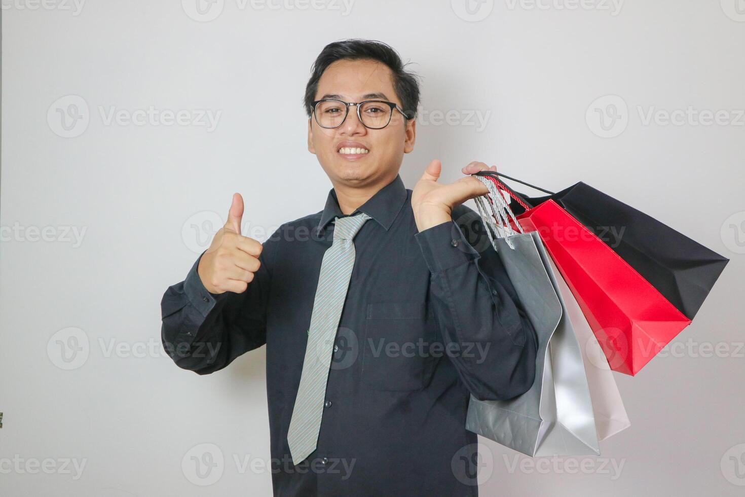 smiling and funny young Asian man in his 30s in black t-shirt standing holding shopping bags with groceries after shopping while showing thumbs up isolated on pastel white background studio portrait. photo