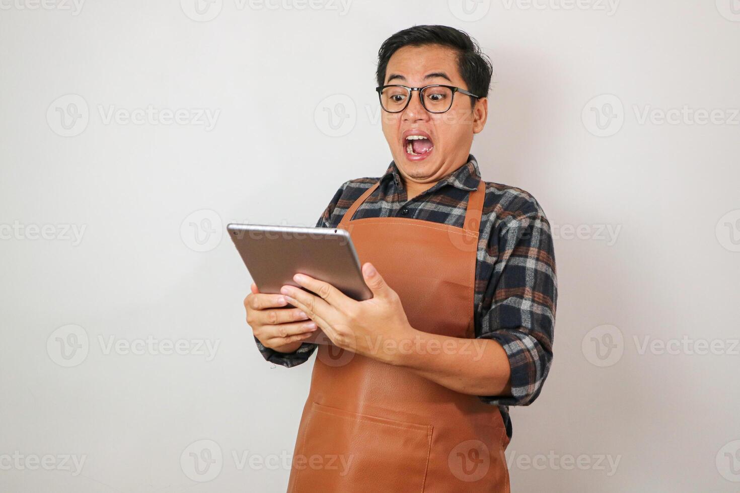 Asian man wearing apron holding tablet with surprised expression looking at tablet, coffee and restaurant business idea concept photo