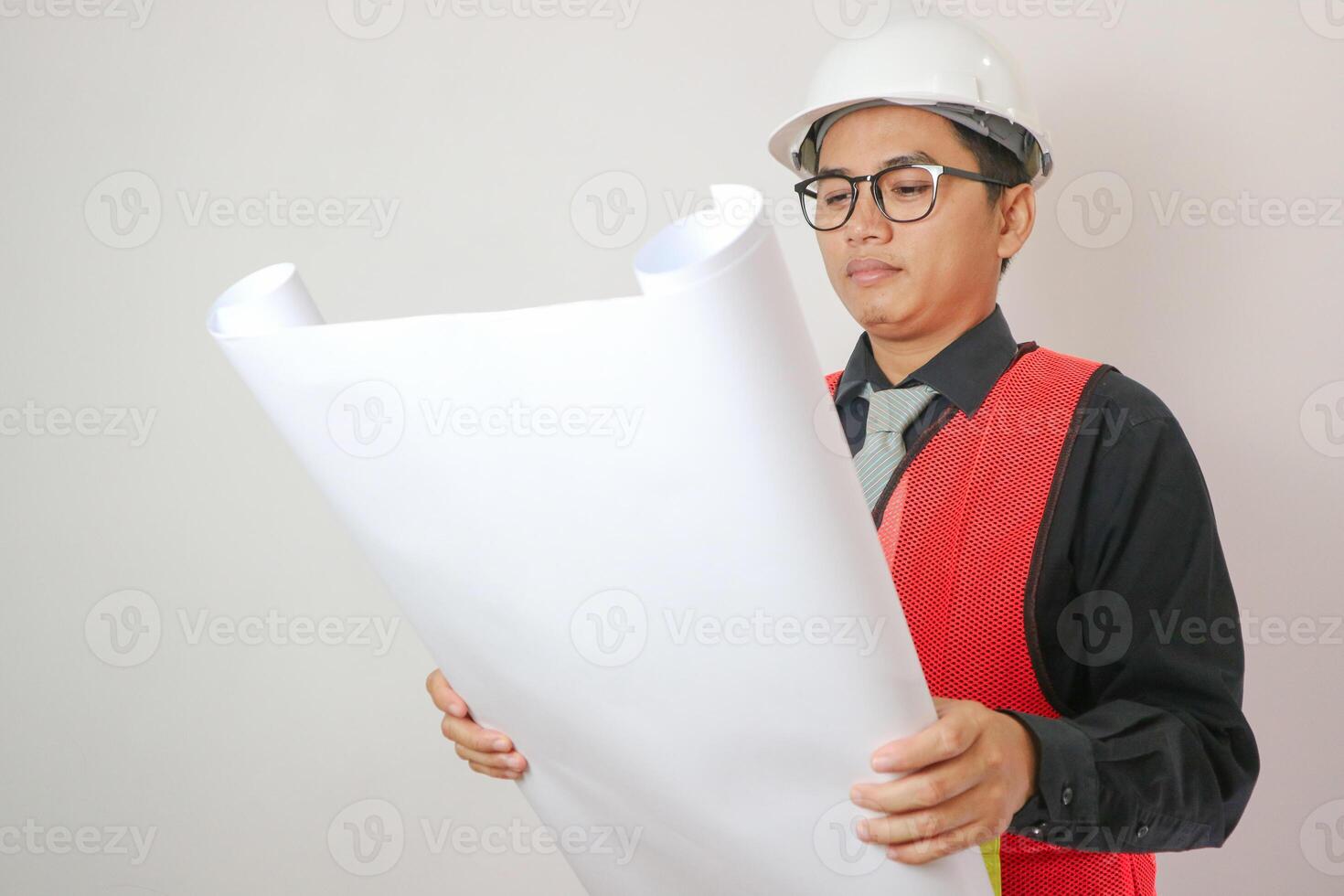 Asian engineer handsome man or architect looking forward holding paperwork blueprint with white safety helmet in construction site. Standing at modern building. photo