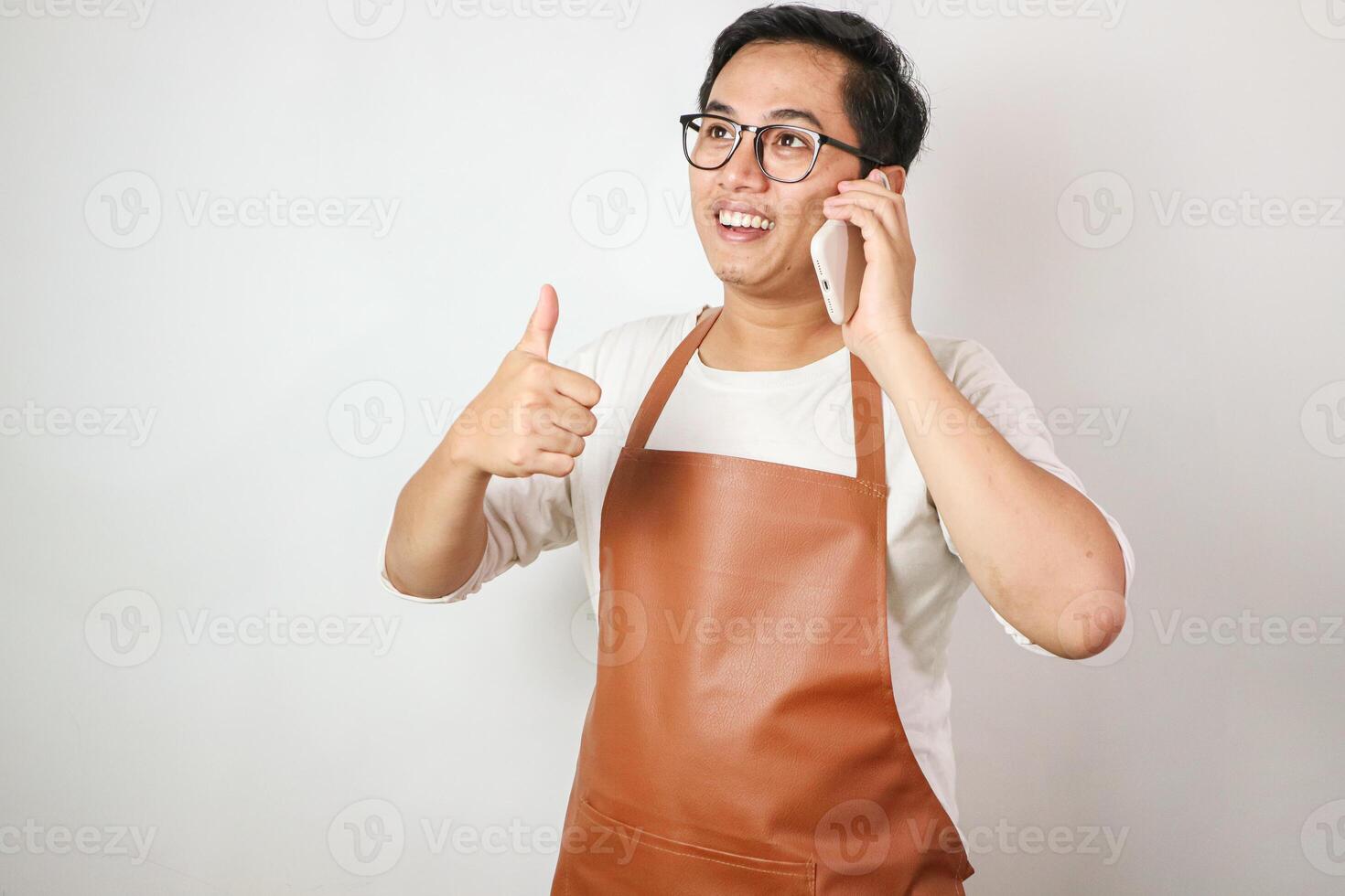 Smiling young Asian man barista barman employee wearing brown apron work in coffee shop talk on mobile phone photo