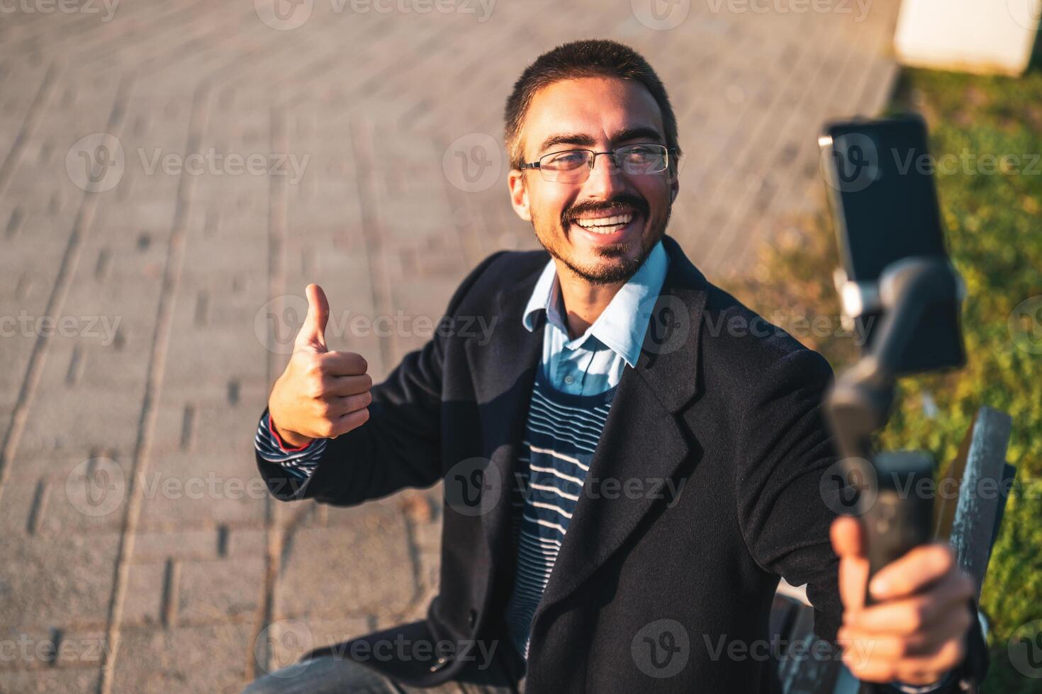 Cheerful businessman vlogger wearing eyeglasses and showing thumb up while using mobile phone with gimbal. He is sitting on a bench and recording vlog outdoor. photo