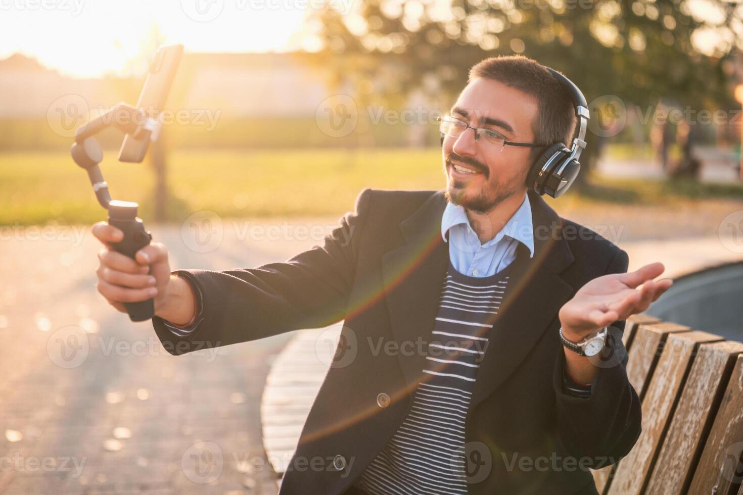 Businessman vlogger in coat wearing headphones and eyeglasses. He is using mobile phone with gimbal in the park and talking while recording vlog on a sunny autumn afternoon. photo
