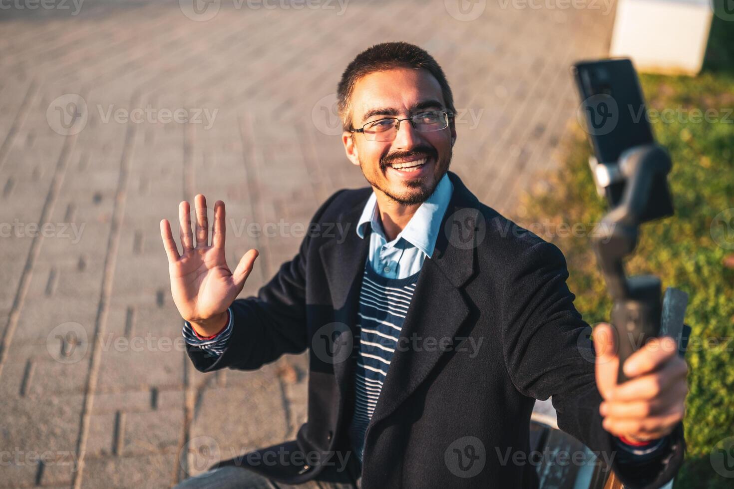 Cheerful businessman vlogger wearing eyeglasses and waving while using mobile phone with gimbal. He is sitting on a bench and recording vlog outdoor. photo