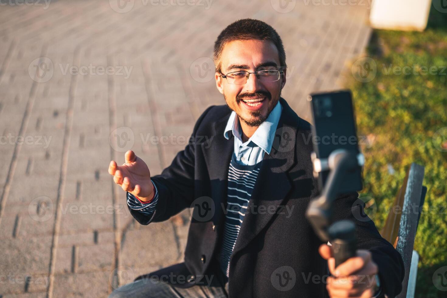 Businessman vlogger wearing eyeglasses and using mobile phone with gimbal. He is talking while recording vlog outdoor. photo