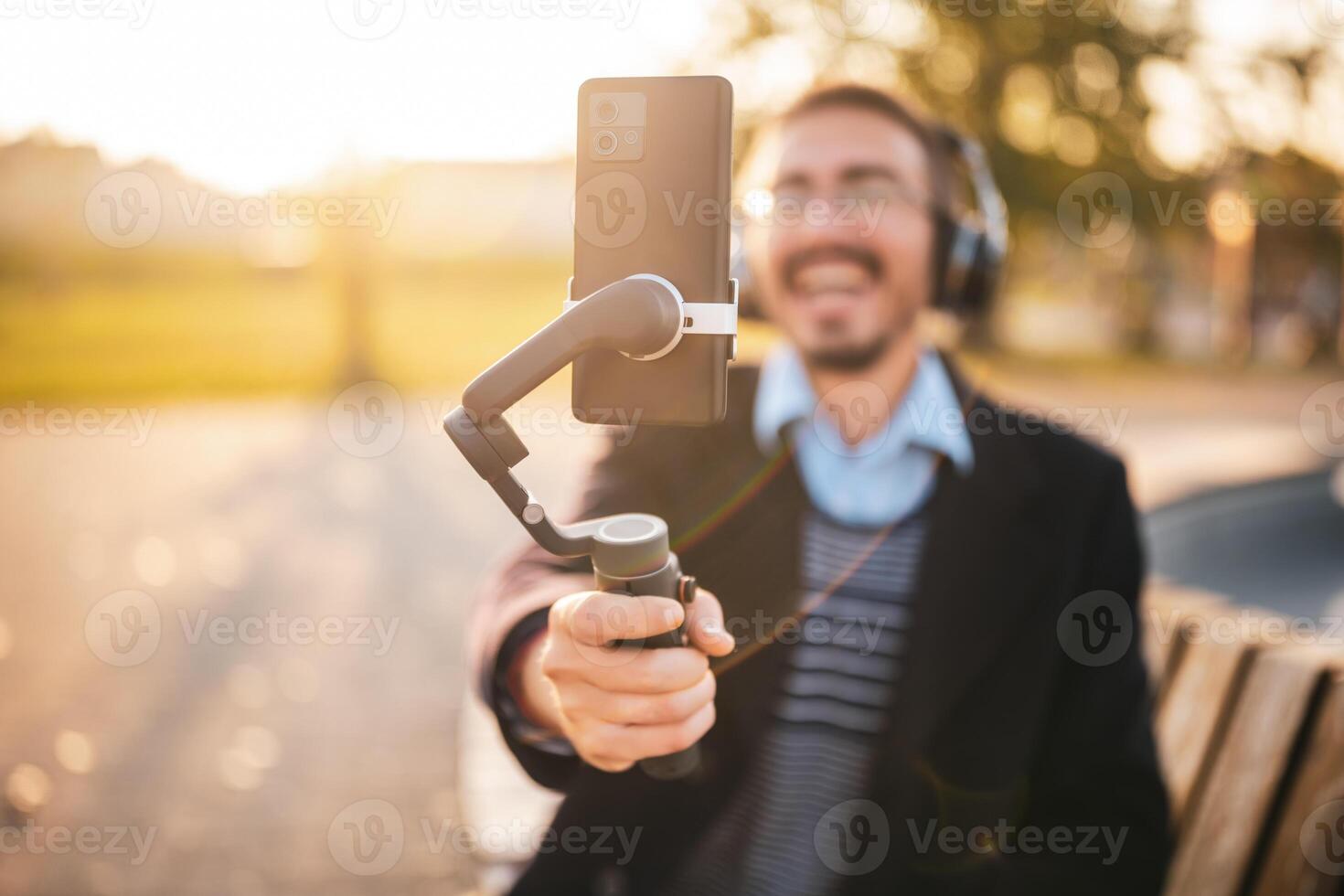 Businessman vlogger is using mobile phone with gimbal while sitting outdoor and recording vlog on a sunny autumn afternoon. He is wearing headphones and eyeglasses. Focus on the phone and gimbal. photo