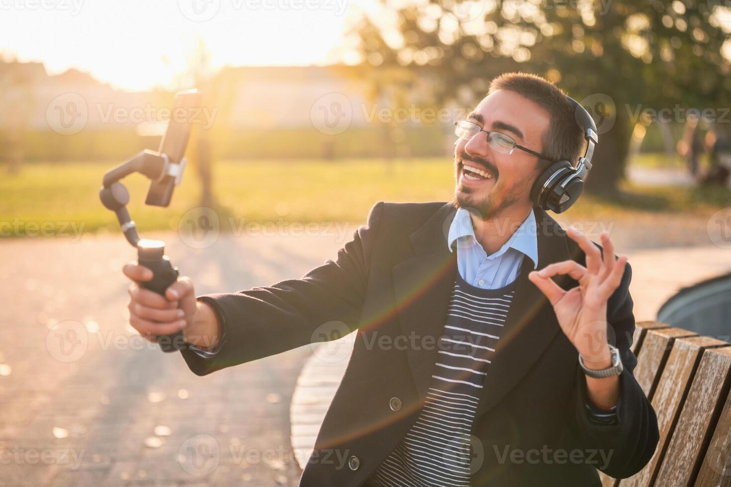 Businessman vlogger in coat wearing headphones and eyeglasses. He is showing ok sign and using mobile phone with gimbal while recording vlog on a sunny autumn afternoon in the park. photo