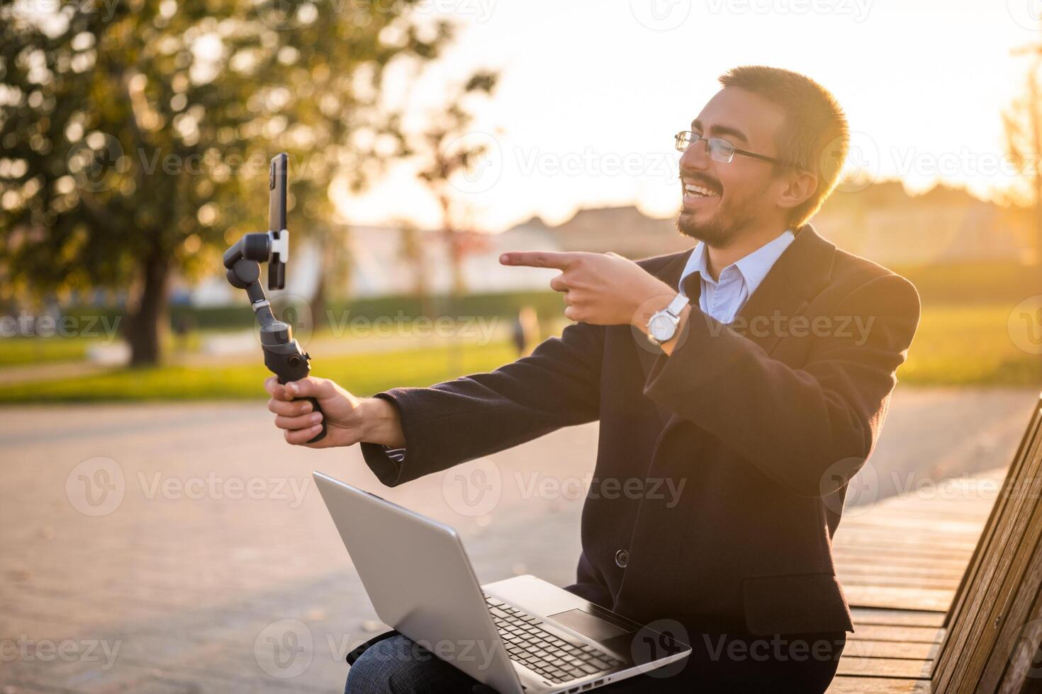 Cheerful businessman vlogger in coat wearing eyeglasses and pointing at mobile phone with gimbal. He is sitting on a bench in the park and recording vlog on a sunny autumn afternoon. photo