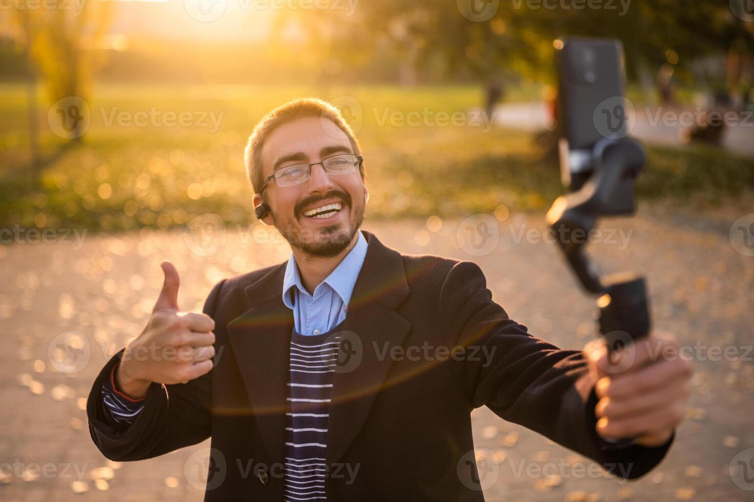Businessman vlogger in coat wearing in-ear headphones and eyeglasses showing thumb up while using mobile phone with gimbal. He is standing in the park and recording vlog on a sunny autumn afternoon. photo