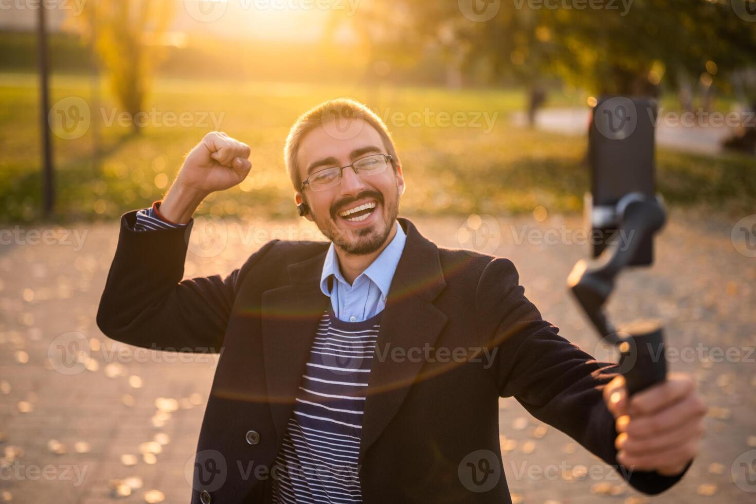Excited businessman vlogger in coat wearing in-ear headphones and eyeglasses gesturing while using mobile phone with gimbal. He is standing in the park and recording vlog on a sunny autumn afternoon. photo