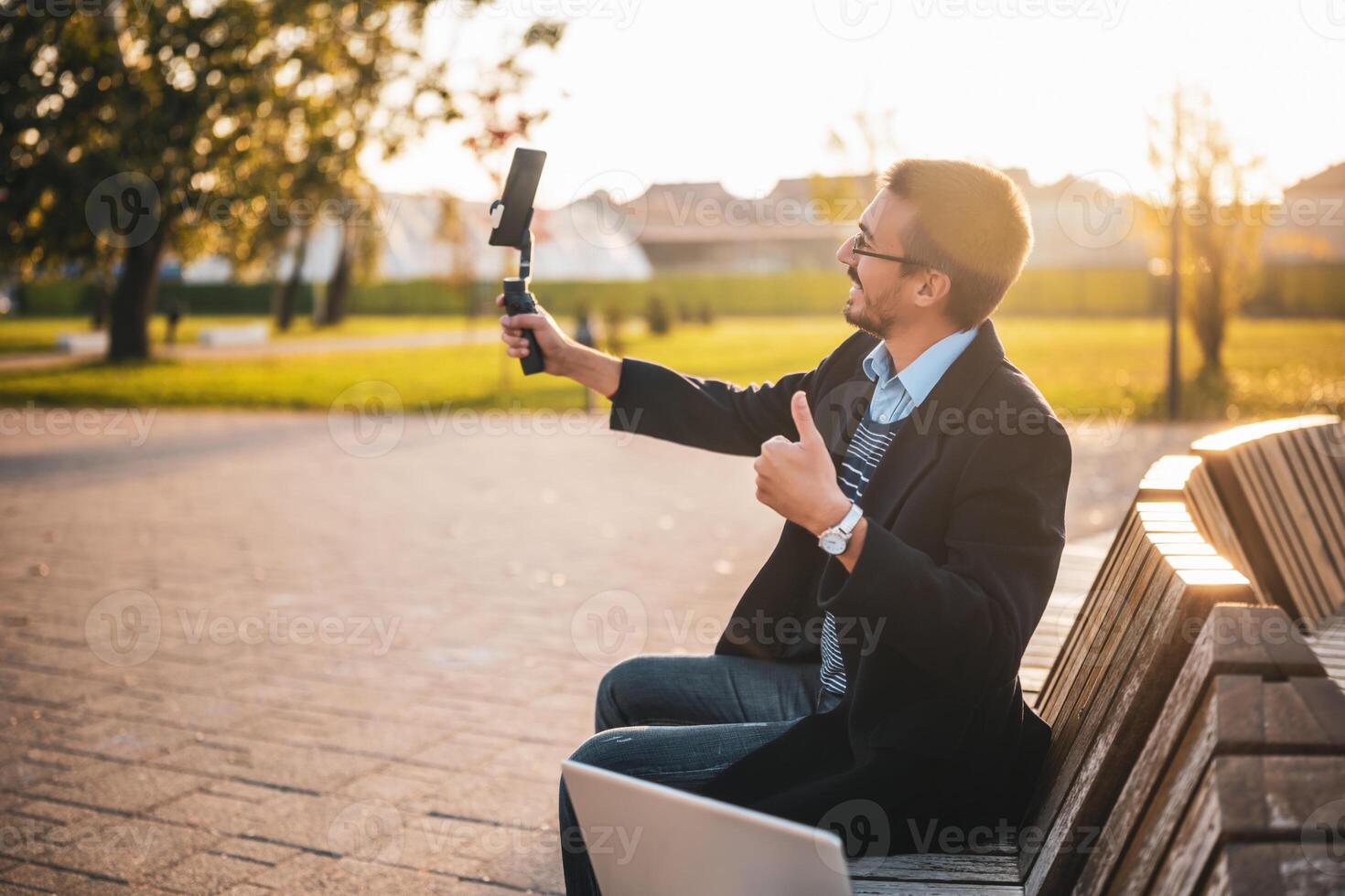 Businessman vlogger in coat wearing eyeglasses and showing thumb up while using mobile phone with gimbal. He is sitting on a bench in the park and recording vlog on a sunny autumn afternoon. photo