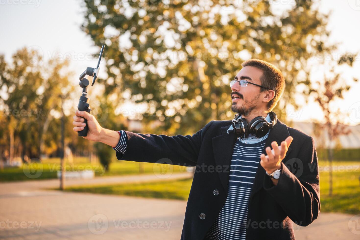 Businessman vlogger in coat wearing eyeglasses and headphones. He is using mobile phone with gimbal in the park and talking while recording vlog on a sunny autumn afternoon. photo
