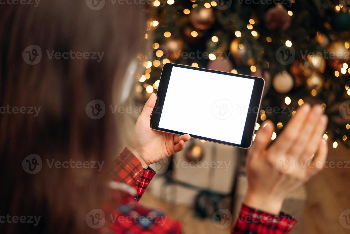 Woman holding tablet with blank screen in front of Christmas tree, festive bokeh lights in background, ready for holiday call or virtual greeting. photo