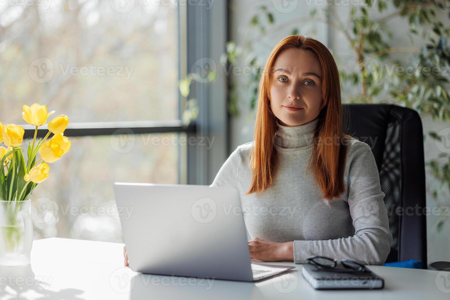 Mid aged woman standing at a desk with a laptop. Cheerful business lady with looking in camera. Happy woman in her workspace, managing a business or working. photo