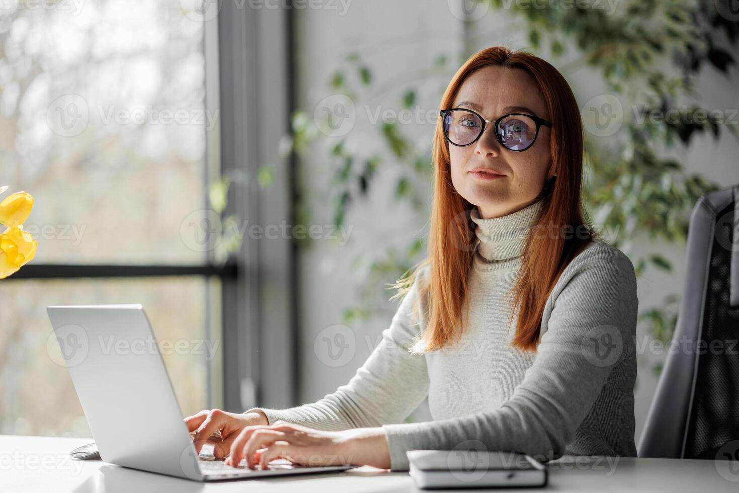 Senior chic woman using laptop at home while taking notes in notebook. Old freelancer works in the living room on a laptop while drafting details on a book. photo