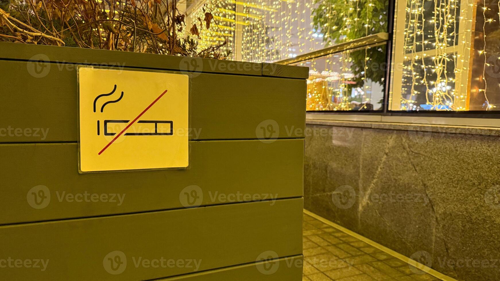 Sign on wooden flower bed against background of glass building with light prohibiting smoking at entrance to house. Prohibited area from smoking cigarettes with special ban sign photo