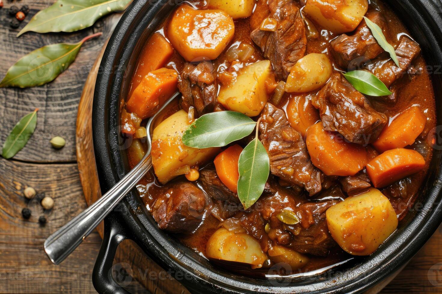 Top view of a black casserole pot filled with a rich beef stew, featuring potatoes and carrots in savory gravy, garnished with bay leaves and served with a spoon photo