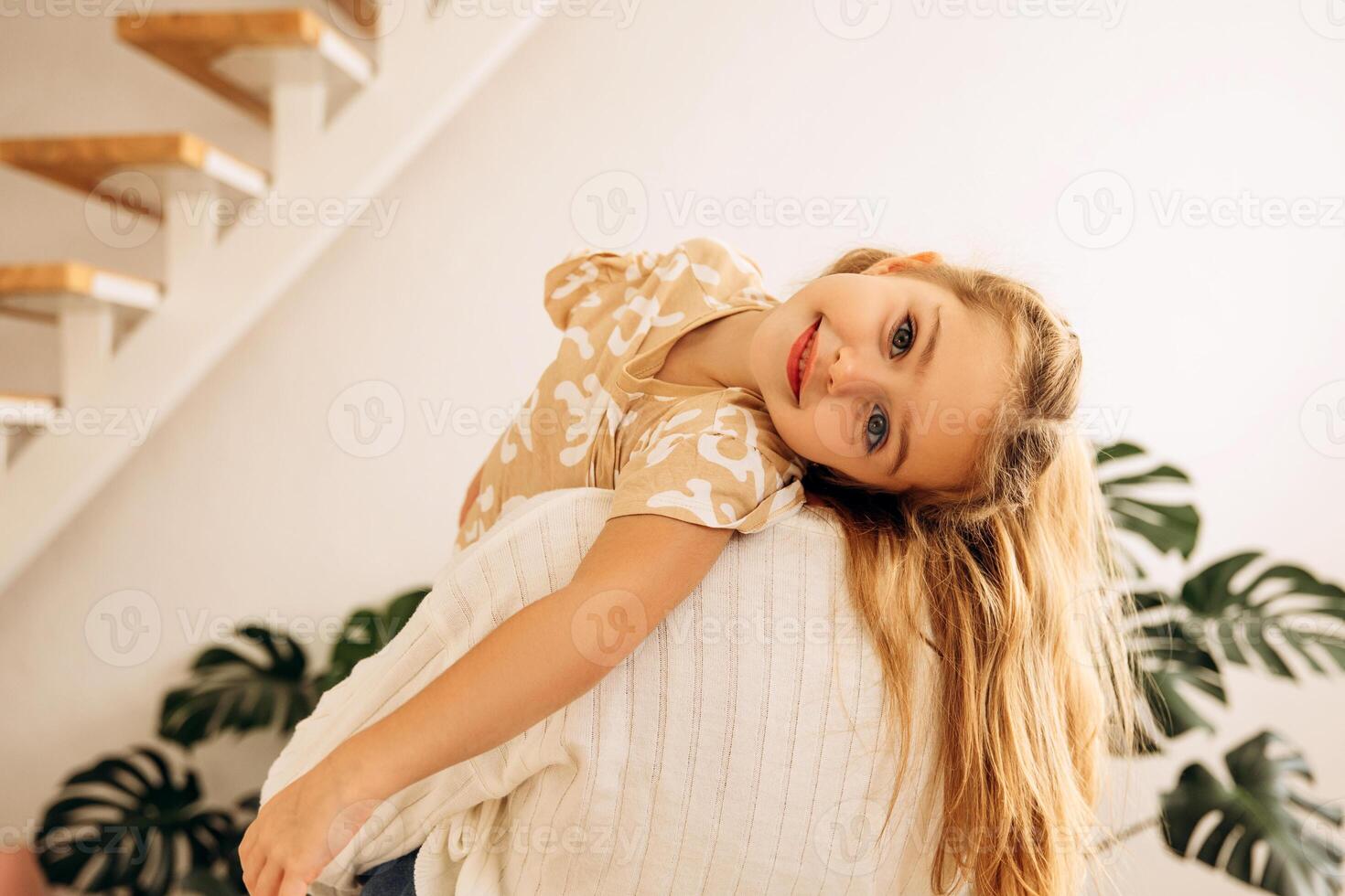 Smiling, beautiful little girl looking at camera, hugging with mother at home in living room photo