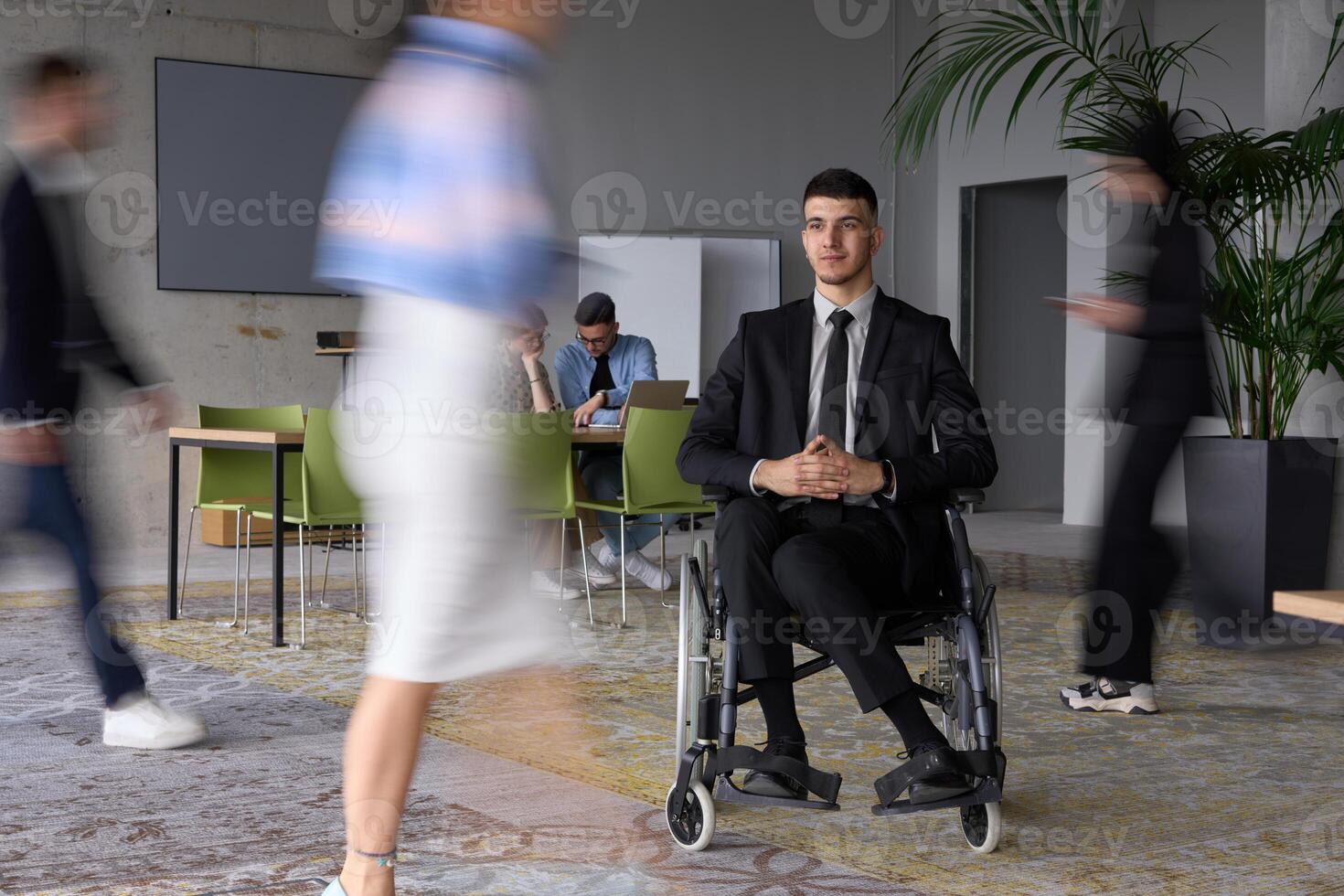 A businessman in a wheelchair navigating through a busy office, surrounded by his colleagues who are actively engaged in their work and collaboration photo