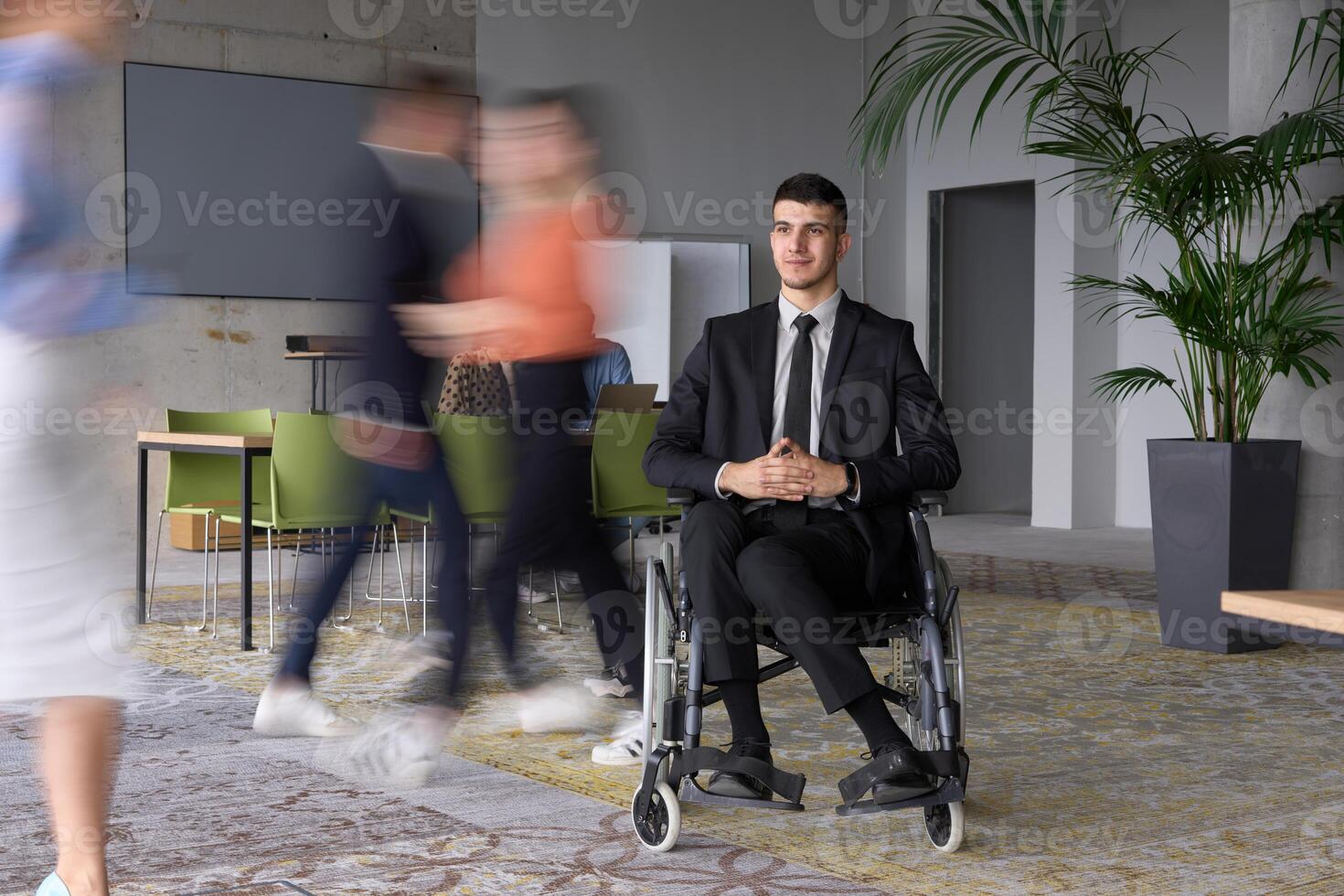 A businessman in a wheelchair navigating through a busy office, surrounded by his colleagues who are actively engaged in their work and collaboration photo